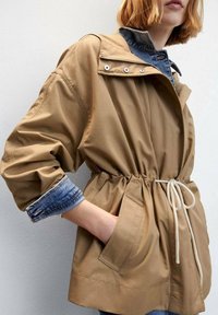 Henkilö, joka on pukeutunut vaaleanruskeaan naruhuppariin denimpaitojen kanssa, joiden hihat on kääritty ylös, seisoo vaaleaa taustaa vasten.