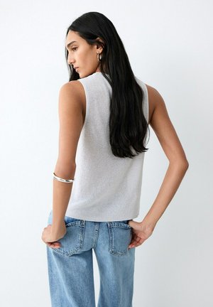 Frau mit langem dunklem Haar, die ein ärmelloses weißes Oberteil, blaue Jeans, silberne Creolen und ein silbernes Armband trägt, posiert mit den Händen in den Gesäßtaschen.