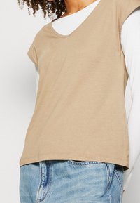 Top beige a maniche corte con scollatura a V, realizzato in tessuto morbido; indossato sopra una camicia bianca a maniche lunghe e abbinato a jeans azzurri chiari.