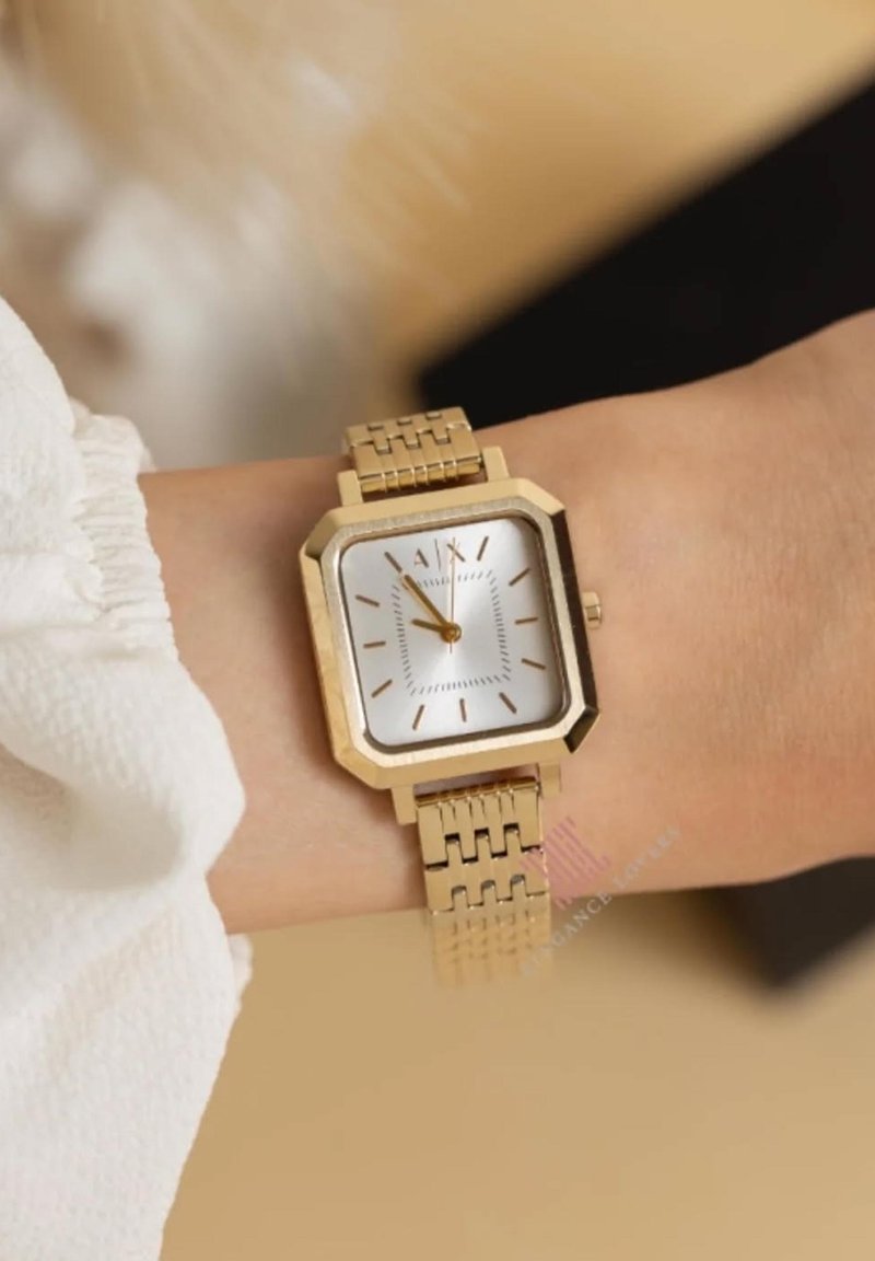 Reloj de pulsera cuadrado en tono dorado con una correa de metal articulada, llevado en una muñeca con una blusa de manga larga blanca y volante.