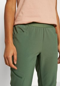 Groene stretchbroek met elastische tailleband, voorzien van zijzakken en subtiele textuurdetails. G gedragen met een lichtroze top.