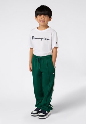 Un jeune garçon souriant se tient avec les mains dans les poches, portant un t-shirt blanc Champion, un pantalon de survêtement vert et des baskets noir et blanc.