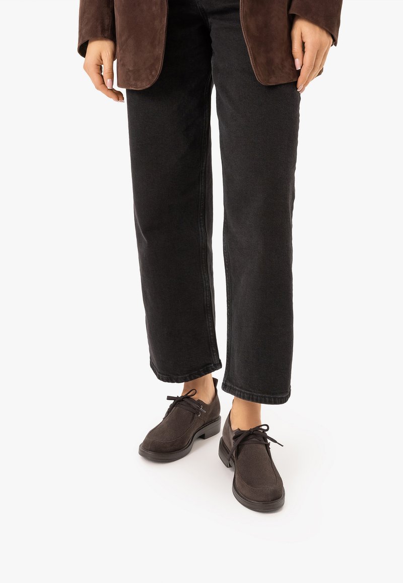 Personne portant un jean noir à jambes droites, des chaussures en daim marron avec des lacets et une veste en daim marron, debout sur un fond blanc.