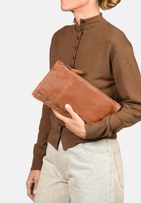 Bolso de mano de cuero marrón con cierre de cremallera, detalles de tachuelas laterales y una textura suave, sostenido por una persona que lleva una camisa marrón.