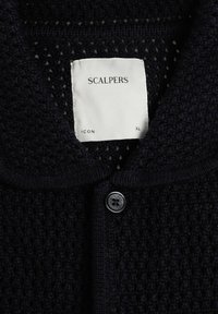 Cardigan noir en tricot avec un motif texturé, col, un bouton noir unique et une étiquette blanche avec l'inscription "SCALPERS ICON XL" à l'intérieur du col.