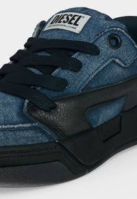 Gros plan d’une basket Diesel en denim bleu et cuir noir avec des lacets noirs et le logo Diesel sur la languette.