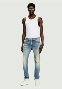 Slim oseba z rumenimi sončnimi očali, belim brezrokavnikom, izblujenimi strganimi modrimi kavbojkami in belimi masivnimi supergami, ki stoji ob enobarvni podlagi.