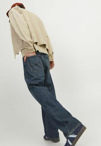 Chaqueta oversized beige con bordes sin rematar, combinada con jeans azules holgados que tienen bolsillos traseros. Footwear oscuro con rayas blancas completa el look.