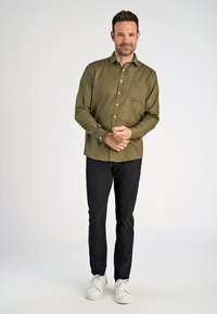 Olivenfarbene Button-up-Bluse mit einer Brusttasche, langen Ärmeln, kombiniert mit schwarzen Hosen und weißen Sneakers vor einem neutralen Hintergrund.