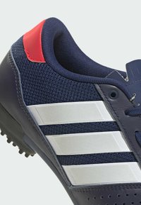 Laivynsininen urheilukenkä, jossa on verkkokangas- ja nahkapäällinen, kolmella valkoisella raidalla, punainen yksityiskohta kannassa ja teksturoitu pohja pidon saavuttamiseksi.