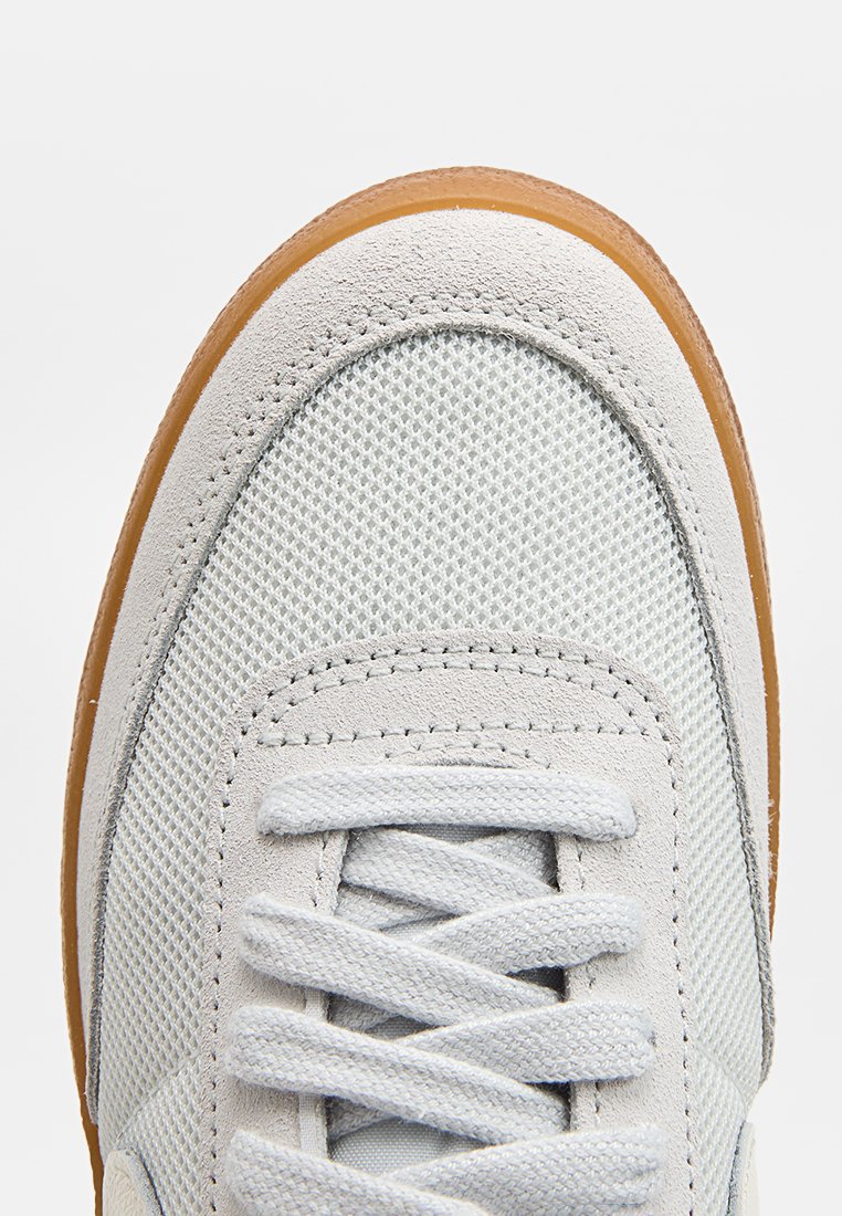Zapatillas gris claro con parte superior de malla texturizada y detalles de ante. Presentan una punta redonda, cierre de cordones y una suela de goma de color caramelos en contraste.