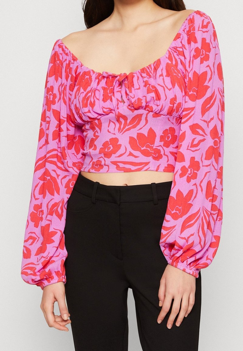 Top court en crochet rose clair à motifs floraux rouges, présentant un design épaules dénudées et des manches longues bouffantes avec des poignets élastiques.