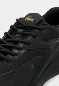 Μαύρο αθλητικό παπούτσι με υφασμάτινες επιφάνειες, χοντρές κορδέλες και χρυσό λογότυπο κροκοδείλου στη γλώσσα, με επενδεδυμένη σόλα.