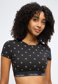 Νεαρή γυναίκα με σγουρά μαλλιά που φοράει ένα μαύρο κοντό τοπ Calvin Klein με λευκά τυπωμένα "cK" και την ένδειξη του бренда στο τελείωμα.