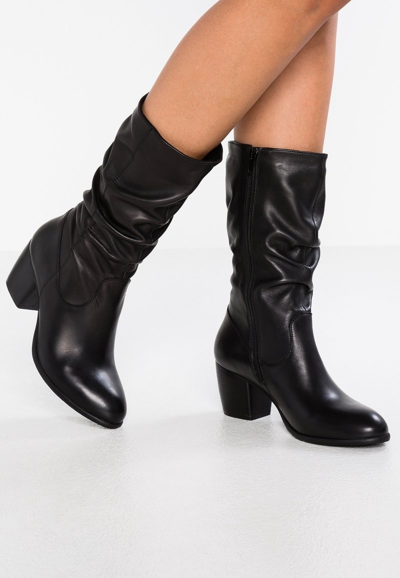 Botas de tacón de cuero negro con un diseño arrugado, puntera afilada y cierre lateral con cremallera. Textura suave con un tacón cuadrado.