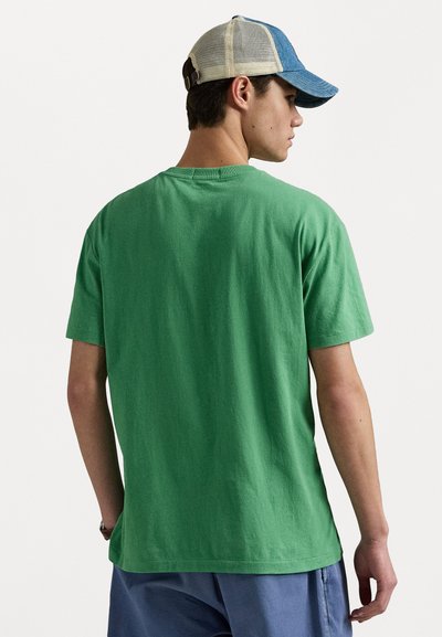 T-shirt en coton vert avec une coupe décontractée et des manches courtes. Le tissu semble doux, avec une texture lisse. Le modèle porte une casquette bleue et beige.