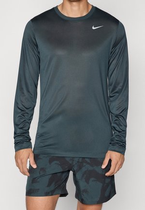 Mężczyzna ubrany w ciemnoszary sportowy long-sleeve Nike i ciemne, wzorzyste krótkie spodenki, stoi z rękami luźno opuszczonymi wzdłuż ciała.