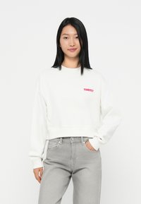 Witte cropped sweatshirt met rode "TOMMY" logo, ronde hals en lange mouwen, gecombineerd met lichtgrijze jeans. Glad materiaal, eenvoudig ontwerp.