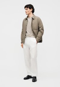 Άνδρας που στέκεται φορώντας μπουφάν ελιάς, ριγέ πουκάμισο σε μπεζ και μαύρο, λευκό παντελόνι και μαύρα επίσημα παπούτσια, με τα χέρια στις τσέπες, στραμμένος προς τα αριστερά.