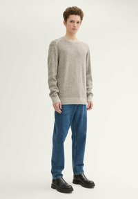 Hellgrauer, strukturierter Strickpullover mit Rundhalsausschnitt, gerippten Bündchen und Saum, kombiniert mit blauen Jeans und schwarzen Stiefeletten, stehend vor einem neutralen Hintergrund.