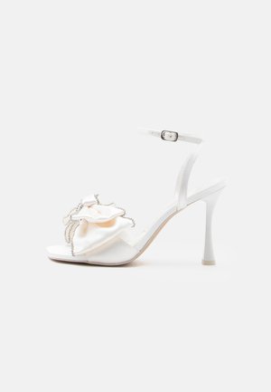 White high-heeled sandal with ankle strap and large decorative fabric flower with rhinestone trim on front strap.