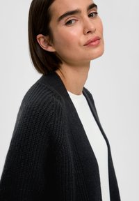 Schwarze Strickjacke mit offenem Design, mit geripptem Muster entlang der Kanten und getragen über einem weißen Oberteil.