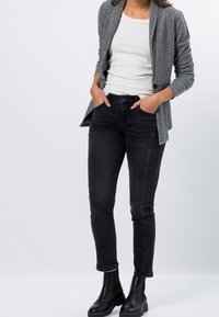 Blazer gris sur un haut blanc, associé à un jean skinny noir et des bottines noires. Tissu lisse, design ajusté et couleurs contrastées.