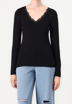 Femme portant un haut noir à manches longues avec un décolleté en V en dentelle et un jean bleu clair déchiré, debout devant un fond blanc.