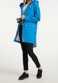 Modrá voděodolná bunda s kapucí, předním zipem, cvoky a bočními kapsami. Nosená přes šedé tričko a černé kalhoty s rovný střih.