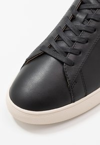 Black leather sneaker with a rounded toe, flat laces, and a cream-colored rubber sole. Stitching accents along the sides.