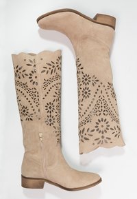 Beżowe zamszowe botki do kolan z wyciętymi wzorami kwiatowymi, jasnobrązowym obcasem i detalem w postaci zamka błyskawicznego z boku, z falbaniastym krawędzią u góry.