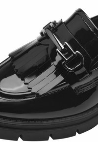 Mocassin en cuir verni noir avec un accent à franges, détails en boucle métallique et semelle épaisse noire. Texture lisse et brillante avec un design élégant.