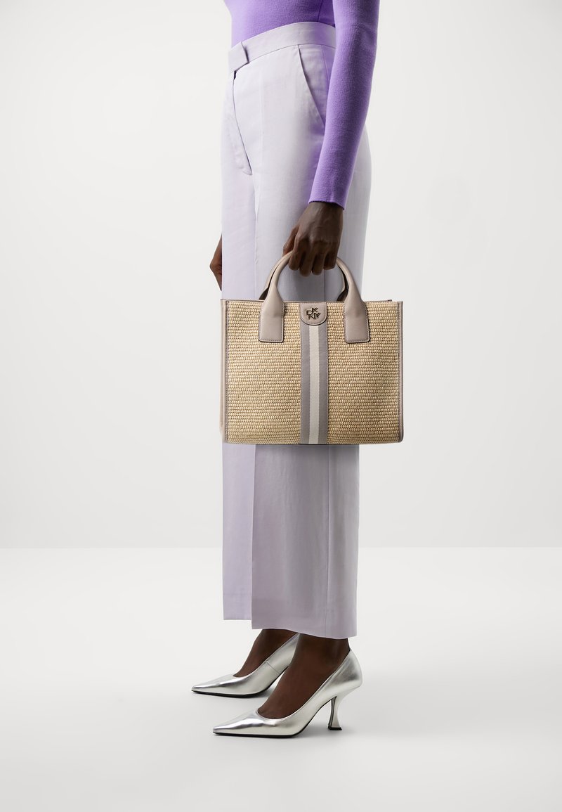 Sac tissé beige avec des rayures grises, des accents en cuir et des poignées supérieures. Le modèle porte un pantalon lilas et des escarpins argentés.