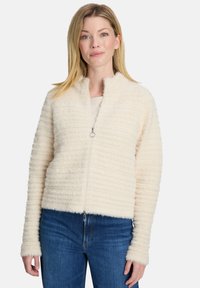 Puszysta kremowa kurtka na zamek błyskawiczny z prążkowaną teksturą, wysokim kołnierzem i długimi rękawami, w zestawieniu z niebieskimi dżinsami. Zawiera srebrny zamek.