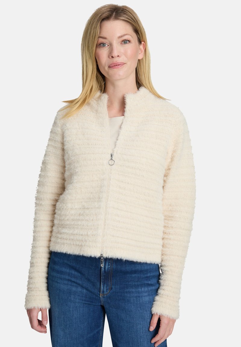 Puszysta kremowa kurtka na zamek błyskawiczny z prążkowaną teksturą, wysokim kołnierzem i długimi rękawami, w zestawieniu z niebieskimi dżinsami. Zawiera srebrny zamek.