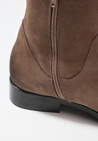 Brązowy zamszowy botek do kostki z bocznym zamkiem błyskawicznym, płaską czarną podeszwą i przeszyciami wzdłuż szwu. Teksturowane wykończenie na górnym materiale.