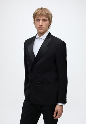 Blonder Mann trägt ein schwarzes zweireihiges Sakko mit Satinrevers über einem weißen Hemd und steht vor einem schlichten Hintergrund.