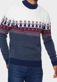 Pull en tricot présentant un haut blanc avec des sections à motifs bleu marine et rouge, incluant des arbres et des motifs géométriques, avec un col côtelé bleu marine.