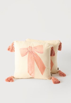 Deux coussins carrés couleur crème avec de grands nœuds roses imprimés à l'avant et des pompons roses à chaque coin sur un fond uni.
