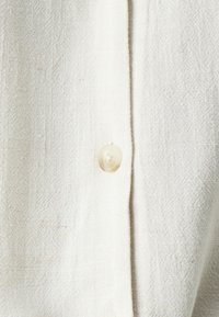 Camicia in lino color crema con una finitura testurizzata, dotata di un singolo bottone in plastica trasparente sulla patta.