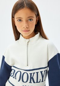 Sudadera con cremallera en blanco y marino con cuello alto y un atrevido gráfico de "BROOKLYN", hecha de tela suave y con un corte relajado.