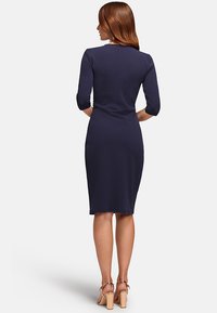 Femme aux cheveux roux portant une robe bleu marine à mi-genoux avec des manches trois-quarts et des sandales à talons beiges, se tenant de dos.