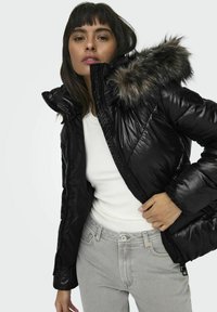 Chaqueta de plumas negra con textura brillante, que presenta una capucha con forro de piel, cremallera frontal y puños elásticos. Lleva puesta una parte superior blanca y jeans grises.