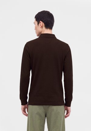 Man met de rug naar de camera gekeerd, draagt een donkerbruine poloshirt met lange mouwen en lichtgroene broek tegen een effen lichte achtergrond.