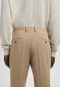 Pantalones beige a medida con frente plano, dos bolsillos traseros y pliegues sutiles, combinados con un suéter de color claro. Textura suave.
