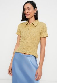 Γυναίκα με κοντά σκούρα μαλλιά, ντυμένη με κίτρινη κοντομάνικη πόλο μπλούζα με υφή και γυαλιστερή μπλε σατέν φούστα, στάση μπροστά σε λευκό φόντο.