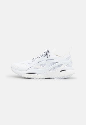 Baskets de sport blanches avec panneaux en maille, forme courbée et semelle rembourrée. Présente un accent noir près de la semelle, des lacets réfléchissants et une branding minimal.