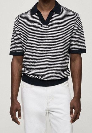 Uomo che indossa una polo a maglia a righe orizzontali bianche e nere a maniche corte e pantaloni bianchi, in piedi davanti a uno sfondo neutro.