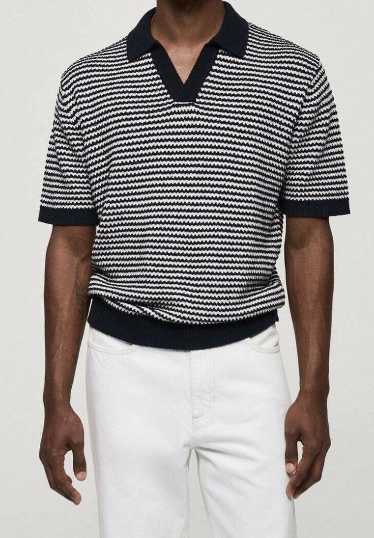 Homme portant un polo en maille à manches courtes rayé horizontalement noir et blanc, et un pantalon blanc, debout devant un fond uni.
