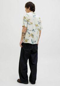 Chemise à manches courtes à motifs floraux avec des couleurs atténuées (crème, vert, bleu), assortie à un jean large en denim sombre et des chaussures marron.